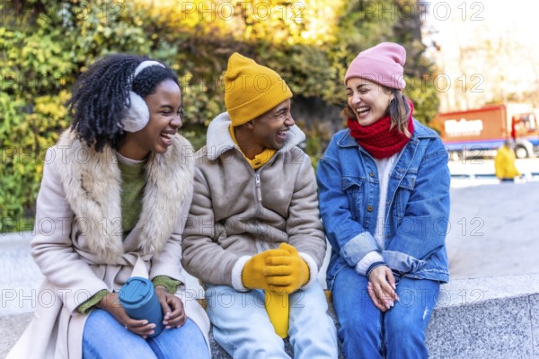 Three cheerful multi ethnic young friends are sitting and laughing together in the city during a winter day