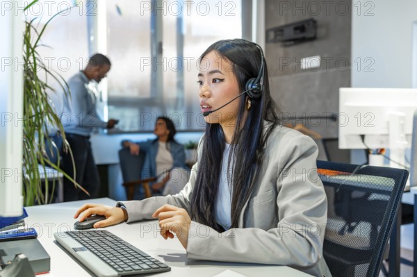Young asian businesswoman wearing headset working with computer in a modern coworking office, colleagues talking in the background