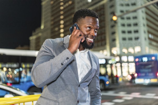 Young businessman smiling while talking on his smartphone in a bustling city street at night, surrounded by vibrant urban lights