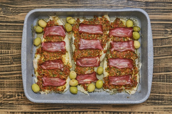 Roasted fish fillets are layered with bacon and herb sauce, accompanied by green olives. This delicious dish is prepared in a baking tray, perfect for sharing