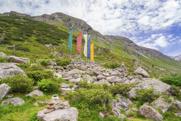 Alpine landscape with colourful flags and a sign next to piles of stones, green vegetation and cloudy sky, Klein Tibet, Zillertal, Austria, Europe