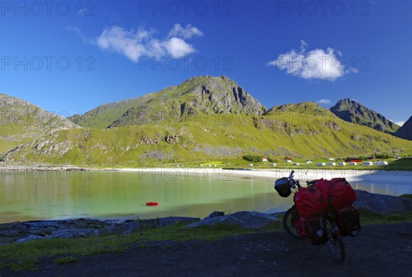 View of a bicycle with red bags on the shore of a bay, surrounded by green hills and mountains under a bright blue sky, bike tours, adventure holidays, Haukland, Leknes, vestavagoya, Nordland, Lofoten, Norway, Europe