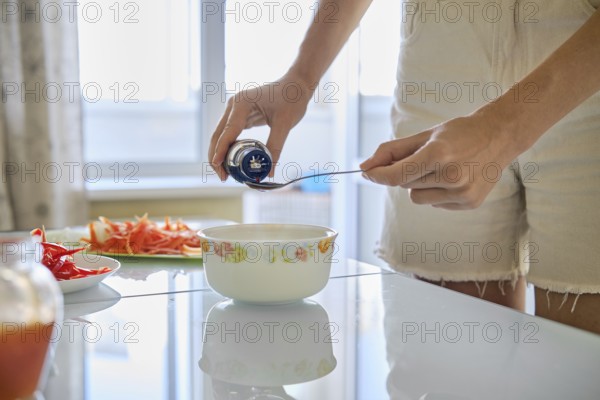An unrecognizable woman adds soy sauce to a bowl while preparing a nutritious meal in a well-lit kitchen. Fresh vegetables are neatly arranged on the counter, indicating a focus on healthy cooking