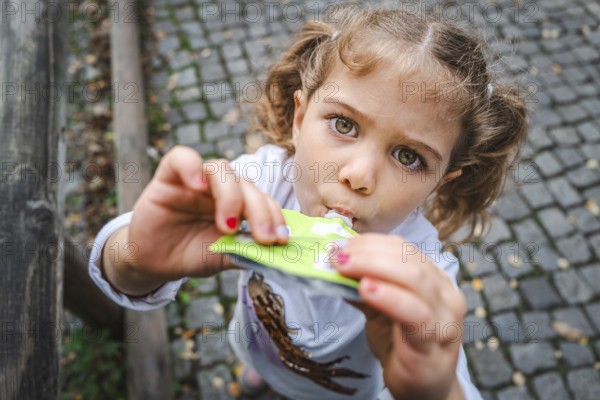 A young girl with pigtails eats a snack on a cobbled street and looks curiously, Stuttgart, Baden-Württmeberg, Germany, Europe