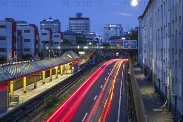Essen, North Rhine-Westphalia, Germany, A40 motorway in the city centre at dusk. Deserted Savignystrasse underground station. On the right, residential buildings behind a noise barrier. On the wall is a LANUV air monitoring station, Essen Kruppstrasse 117 (EKRU2), on the right under the lantern. Residents are affected by noise and exhaust fumes. At the back, buildings with Postbank, Eon and Evonik lettering. Moon in the sky, Europe