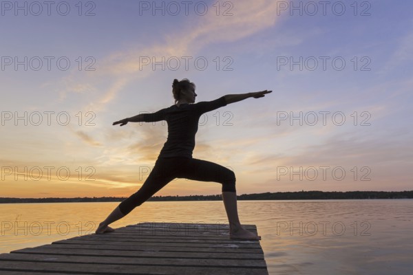 Woman practicing yoga posture Virabhadrasana II, Warrior Pose, lunging standing asana on jetty at lake at sunset
