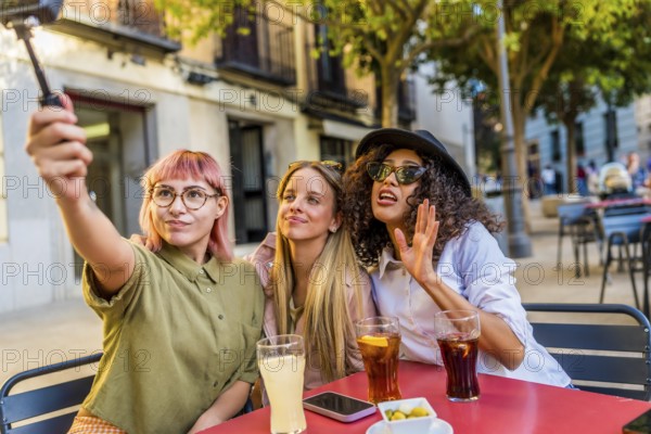 Three modern diverse women taking selfie with digital camera and tripod at the terrace of a bar