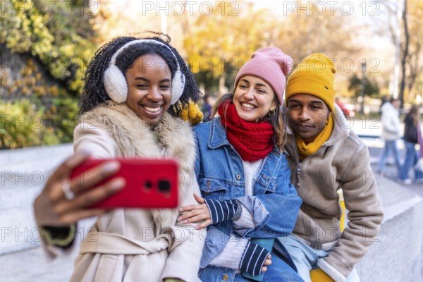 Three happy multi ethnic friends wearing winter clothes taking a selfie with a smartphone in a city park