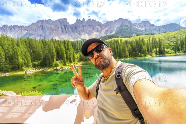 Tourist gesturing peace sign while taking a selfie at lake carezza with latemar mountain range in the background