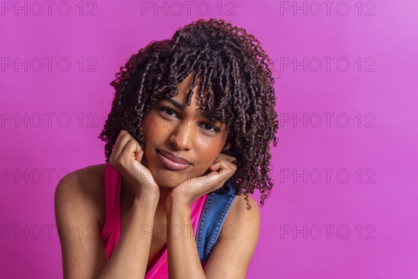Portrait of a thoughtful young woman with curly hair resting her chin on her hands against a vibrant pink backdrop