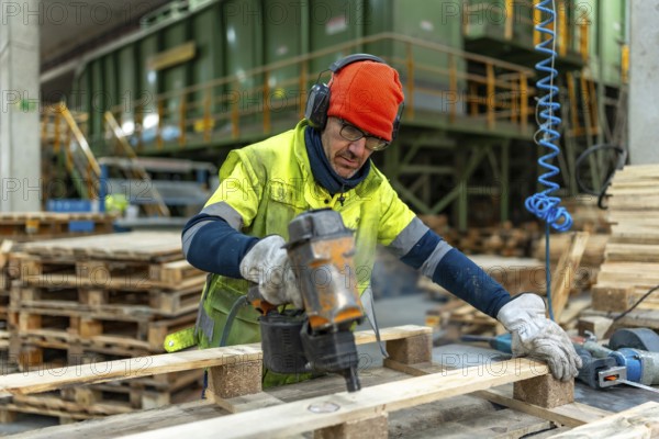 Man with protective gear repairing pallets in a recycling plant warehouse using a compressed air electric machine