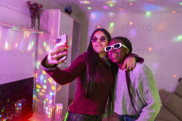 Two teenage girls wearing sunglasses are taking a selfie with a smartphone at a home party with disco lights
