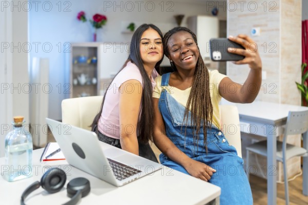 Two happy multi ethnic female students taking a selfie with a smartphone while studying at home