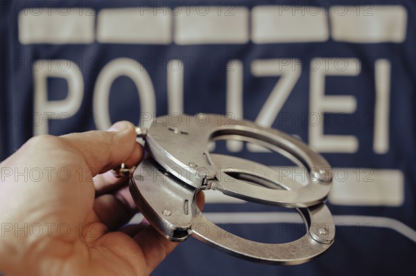 A pair of handcuffs is held by one hand, a police sign in the background