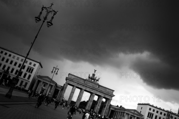 People visit the Brandenburg Gate on a day with a threatening, dramatic sky and heavy clouds, light shines in the background from a gap in the clouds, symbol for Germany, threat, political and economic situation, fear, confidence, hope, early classicism, Triumphtor, archway with quadriga, black and white photo, Pariser Platz, seen from the street Unter den Linden, Berlin Mitte, city of Berlin, Germany, Europe