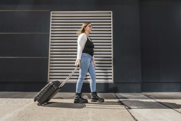 Young business woman pulling rolling luggage along a sunny city sidewalk, walking past a sleek modern building, embodying the essence of urban travel and adventure
