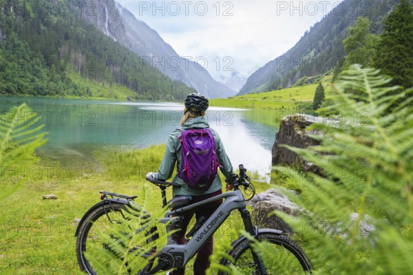 A woman with a helmet and purple rucksack stands next to an e-bike by the lake and looks out over the surrounding mountains, Stilluptal, Zillertal. Austria