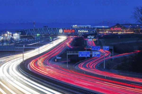 A8 motorway at Stuttgart Airport in the evening. Heavy traffic, lanes of light. Modern architecture of Messe Stuttgart with multi-storey car park above the motorway lanes. Logo of the Bosch company. Stuttgart, Baden-Württemberg, Germany, Europe