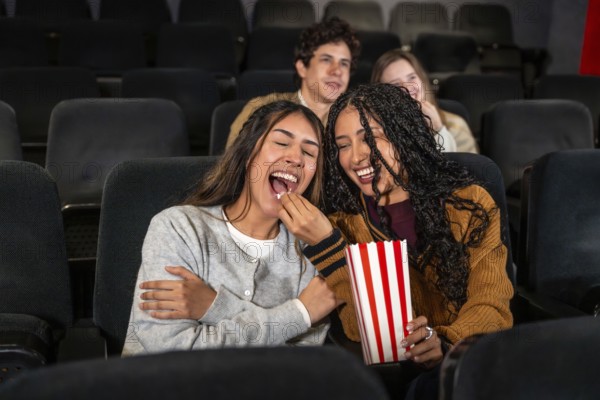 Two young women are enjoying a movie at the cinema, laughing and sharing popcorn, with other people in the background also watching the film