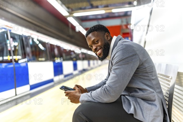 Young black businessman sitting on a bench, using a smartphone while waiting for a subway train at an urban station platform