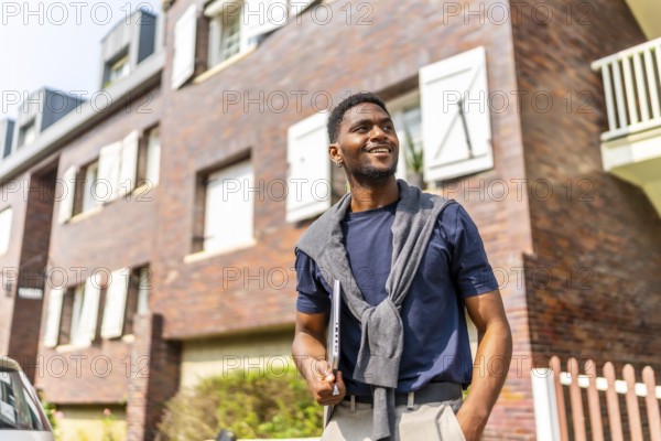 Proud african young businessman with casual clothes carrying laptop looking away standing in the street in a residential area