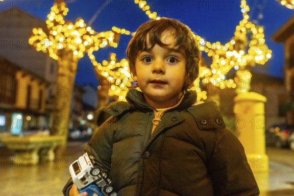 A young boy is standing in front of a Christmas tree with a toy car in his hand. The scene is festive and joyful, with the lights on the tree and the boy's smile