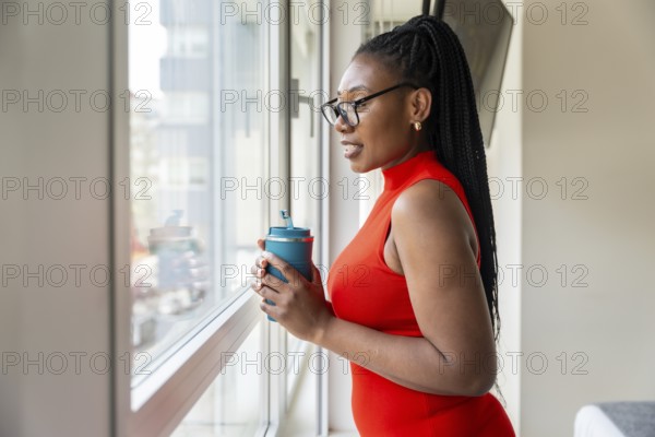 Black businesswoman enjoying a moment of peace while drinking from her travel mug and looking out the window at the cityscape during her business trip