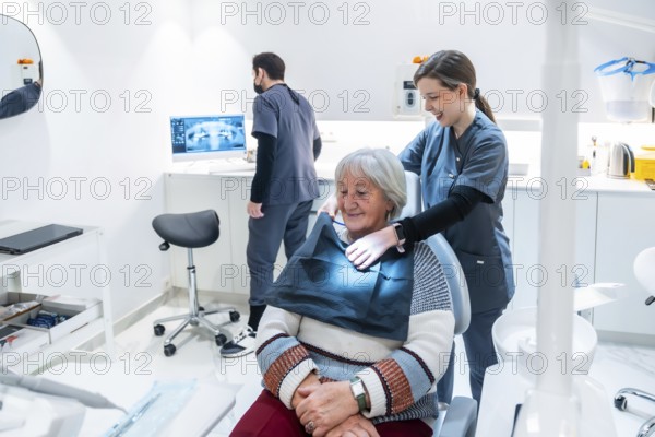 Dentist adjusting bib on senior woman for a dental checkup in a modern clinic, while a technician reviews a radiograph
