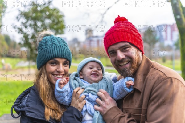 Smiling parents holding their laughing baby wearing winter clothes, enjoying a sunny winter day at the park