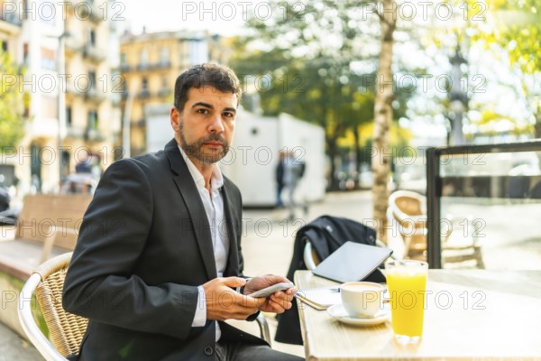 Elegant adult caucasian businessman using phone while drinking coffee in an outdoors cafeteria in the city