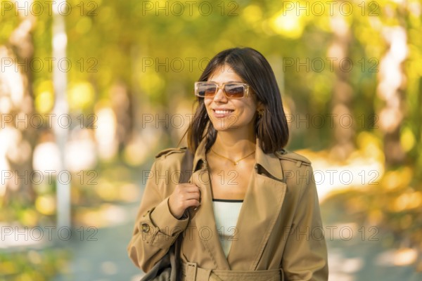 A woman wearing sunglasses and a brown coat is walking down a path. She is smiling and she is enjoying her walk. Autumn or winter concept in the city