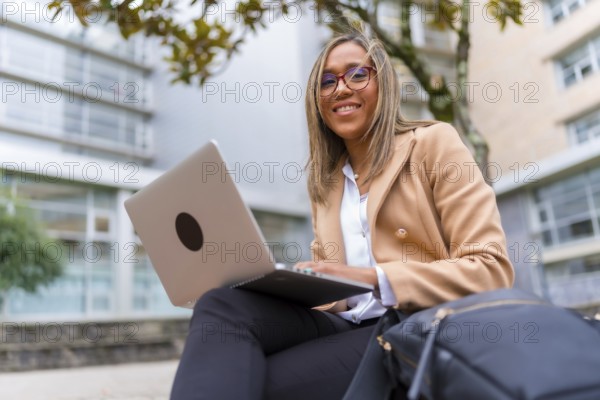 Happy latin businesswoman working with laptop sitting in the city next to a financial building