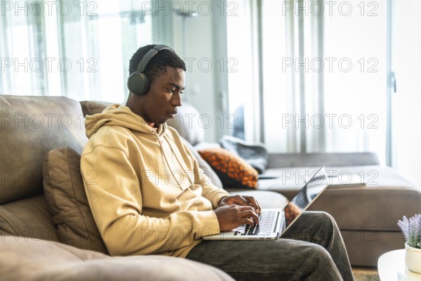 Young man sitting comfortably on a sofa, using a laptop and headphones, focused on studying and working from home in a modern living room