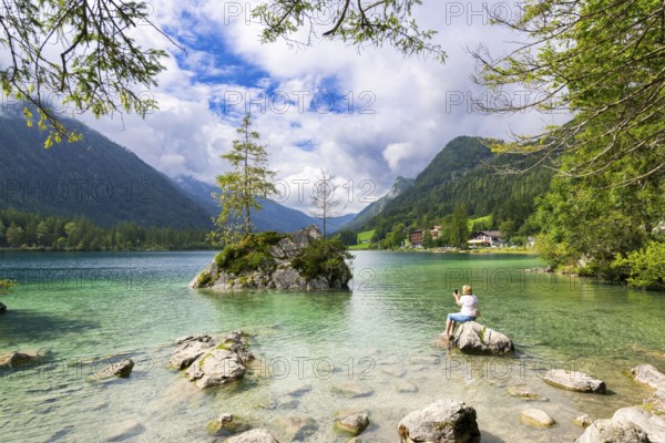 Hintersee with crystal clear water and mountains in the background, a woman sits on a rock and enjoys the picturesque landscape, Berchtesgaden National Park, Berchtesgadener Land, Upper Bavaria, Bavaria, Germany, Europe