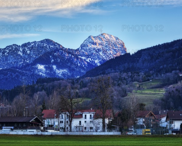 Idyllic village view with snow-covered Mount Wendelstein in the background and a green meadow under a blue sky, Bad Feilnbach