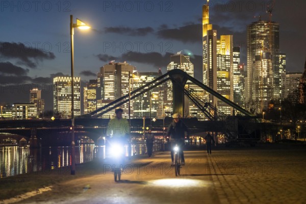 Skyline of the city centre of Frankfurt am Main, cyclist with light on the cycle path, pavement, promenade along the river Main, dusk, Hesse, Germany raft bridge in the background