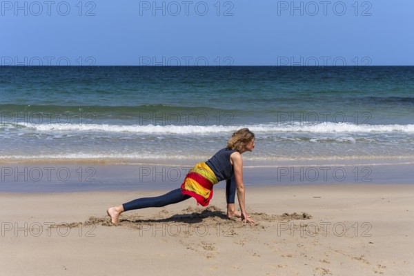 A woman doing yoga on a sandy beach by the sea. Ashwa Sanchalanasana or Sprinter, variant. Treen Cove, Cornwall, England, Great Britain