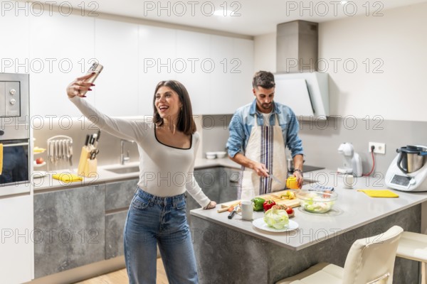 Happy couple preparing healthy meal in their kitchen, woman taking selfie while man is cutting vegetables