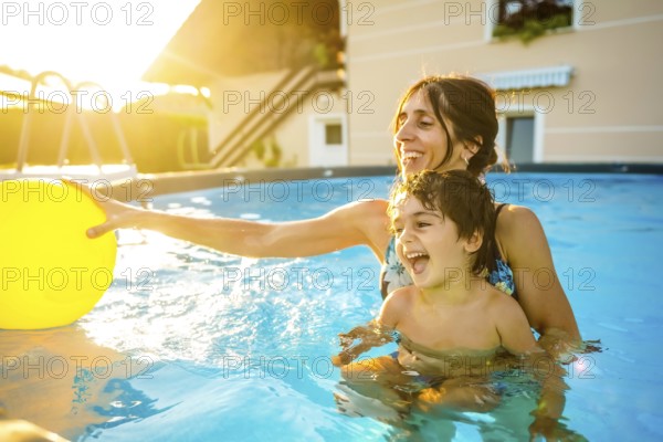 Happy mother and son laughing and playing with a yellow inflatable ball in a home swimming pool during a sunny summer vacation