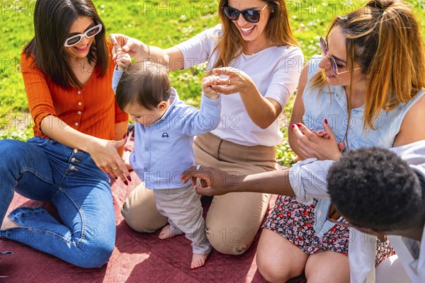 Group of happy multi ethnic friends having fun with a baby learning to walk during a picnic in a park on a sunny day