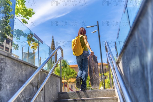 Low angle view photo of the rear view of a young woman with casual denim clothes and a bag carrying a skateboard walking up stairs in the city