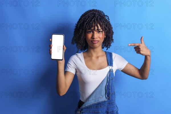 Young woman showing a smartphone with blank white screen and pointing at it with index finger, promoting mobile app or website
