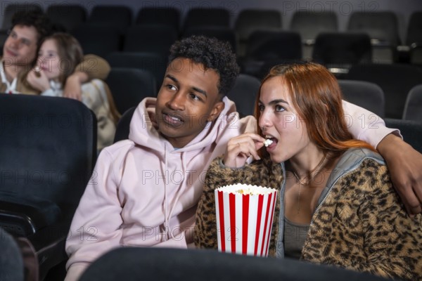 Young multi ethnic couple enjoying a movie and sharing popcorn in a cinema, surrounded by other people