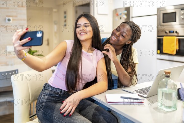 Two happy teenage girls taking a selfie with a smartphone while doing homework in a modern kitchen