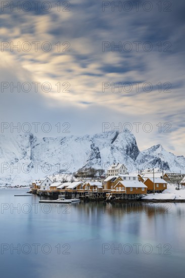 Traditional yellow rorbuer huts on the island of Sakrisøy, snow-capped mountains in the background, Sakrisoy, Reine, Lofoten, Norway, Europe