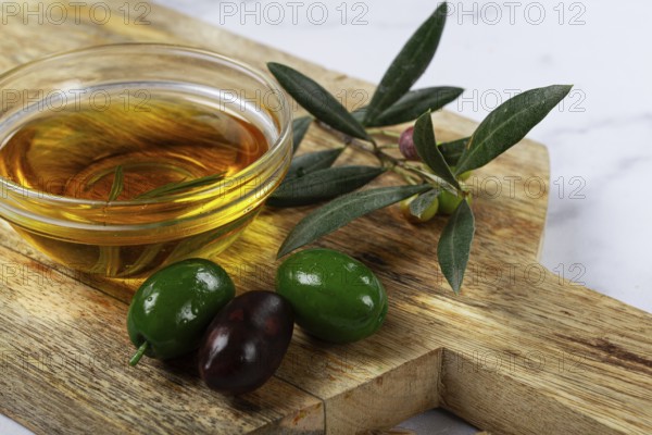 A glass bowl filled with rich olive oil sits on a wooden cutting board alongside fresh green and black olives, complemented by olive leaves, creating a rustic culinary setting