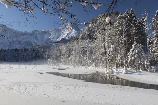 Reflection of trees on Lake Almsee which is partly frozen and snow covered. Snow on the forest with blue sky
