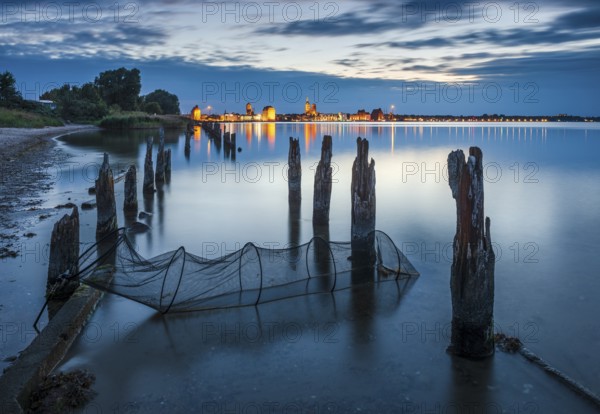 View at dusk over the Strelasund to the illuminated town of Stralsund with its churches and the warehouses in the harbour, in front old groynes and a fishing net, Stralsund, Mecklenburg-Vorpommern, Germany, Europe