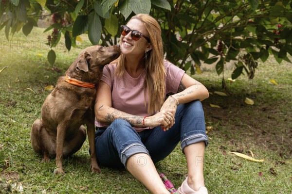 The happy woman with sunglasses and her dog sitting in the park, her pet gives her a kiss on the cheek