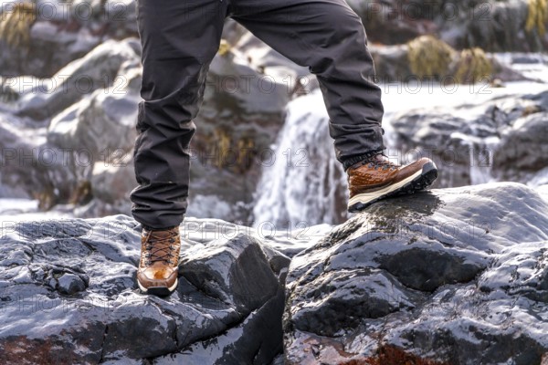 A person is standing on a rock near a waterfall. The person is wearing brown boots and gray pants